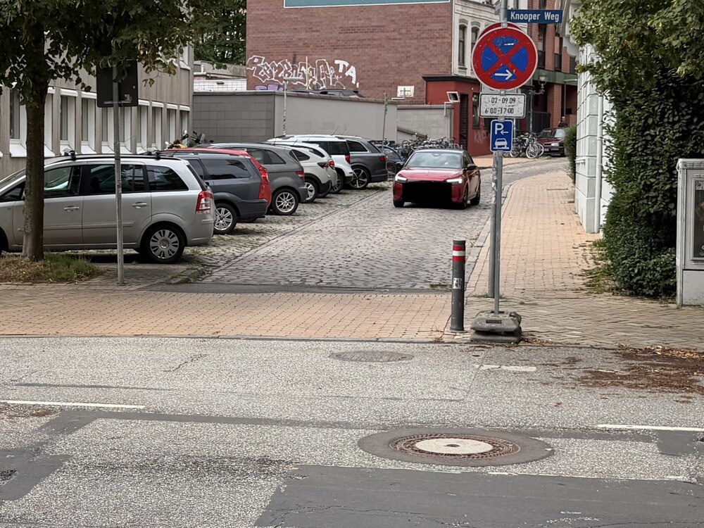 Blick &uuml;ber den Knooper Weg an einer Einm&uuml;ndung: Fahrzeuge stehen l&auml;ngs und schr&auml;g auf einem gepflasterten Streifen neben der Fahrbahn, rechts eine Halterung mit Haltverbot, Zusatzzeichen und blauem Gehwegparken-Schild.
